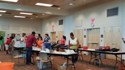 People at tables in a large room, possibly an event or distribution. Boxes and supplies are on the tables.