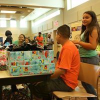 People at a table with gifts. Man in orange shirt, woman with green shirt, boxes, and a school setting.