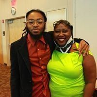 Man and woman smiling, indoors. Man in black blazer and glasses. Woman in green top and glasses.