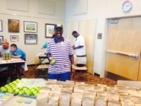 Man in striped shirt at table, surrounded by bags and green items. Other people in the background, indoor setting.