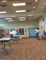 A room with tables, quilts on display, and people. Brown carpet, beige walls, and white balloons.
