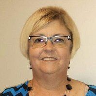 Woman with blonde bob and glasses smiles, wearing a blue patterned top and necklace.