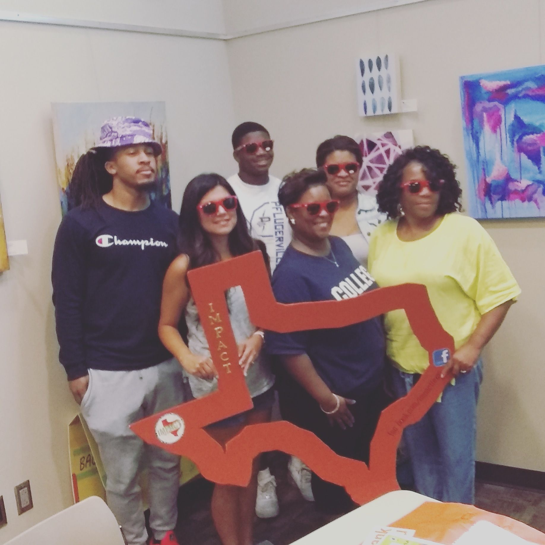 Group of people wearing red sunglasses posing with a red Texas-shaped frame. Artwork on walls.