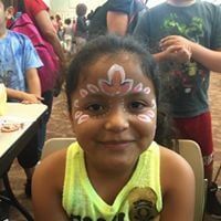 Girl with face paint smiles, sitting in a chair at an event.