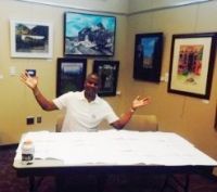 Man sitting at a table with papers, arms raised in a room with framed artwork.