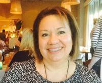 Woman smiling, seated at a restaurant table. Wearing a black and white patterned top. Other people are in the background.