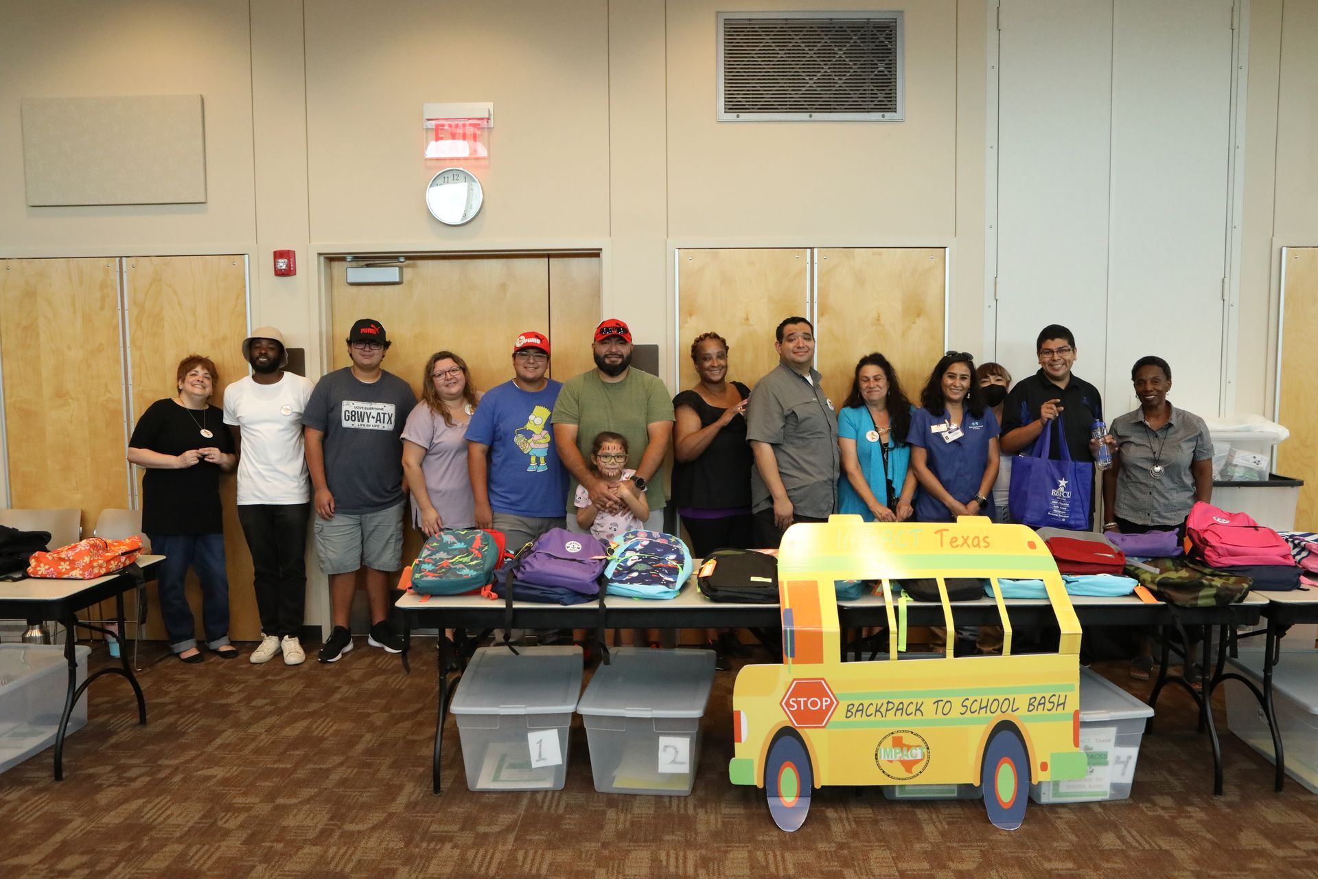 Group of people stand behind tables with backpacks, a school bus cutout, in a room.