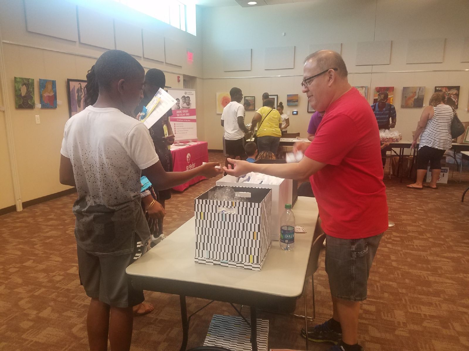 A man in red shirt handing something to a person in a white shirt at a table with a striped gift bag.