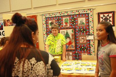 People at a vendor table with a Christmas-themed quilt in the background. Woman in a tie-dye shirt stands behind the table.