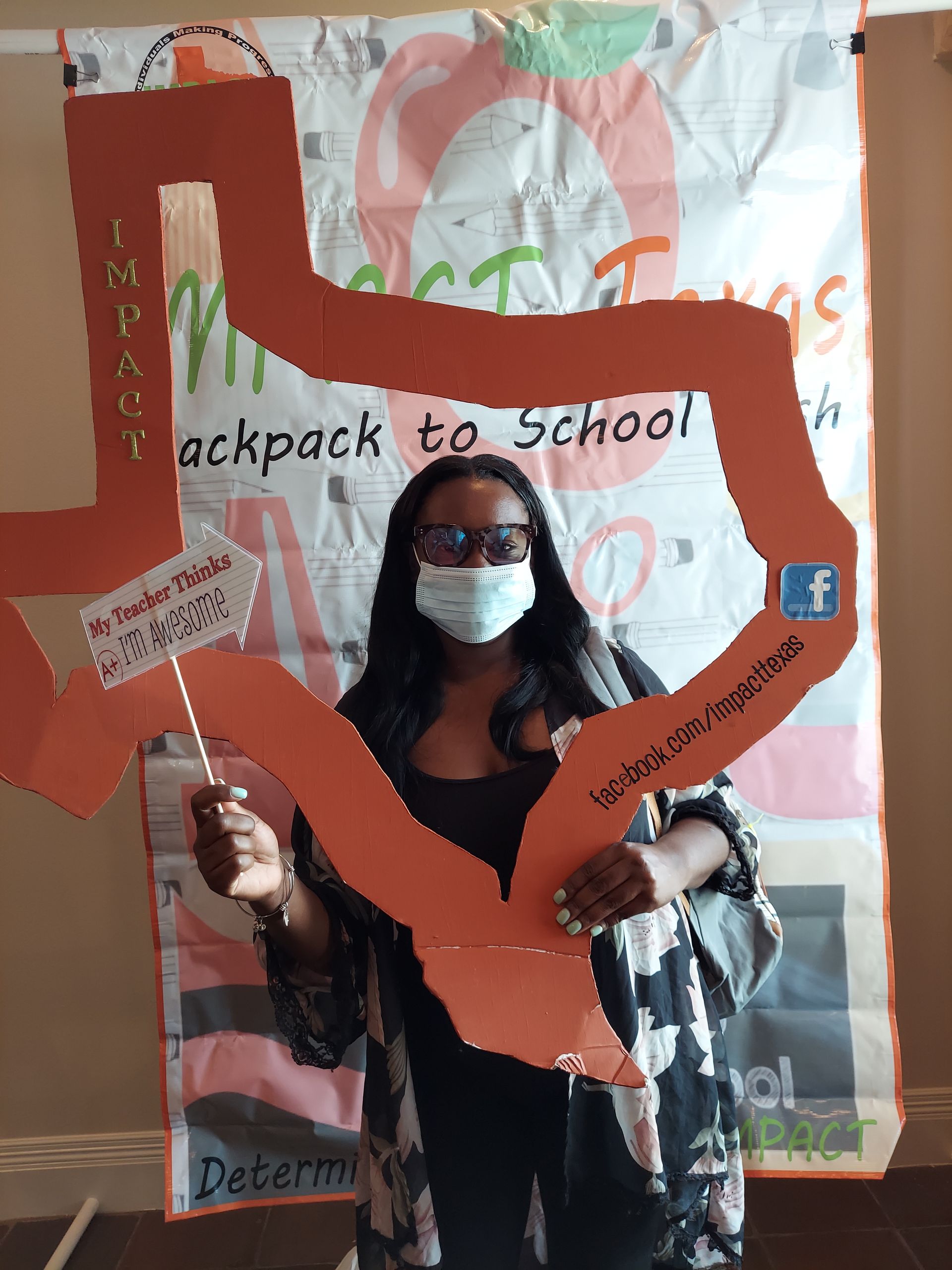 Woman in mask holds an orange Texas-shaped sign, smiling, in front of a back-to-school banner.