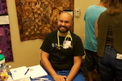 Man with stethoscope, smiling, seated at a table. He wears a black shirt, blue scrubs, and is in a room.