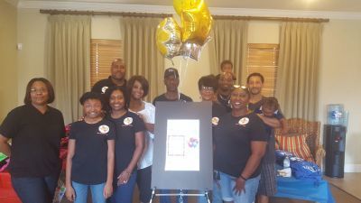 Group of people holding a large framed picture, smiling, with balloons inside a home.