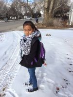 Girl standing in snow, wearing a black coat, scarf, backpack. Snowy road, leafless trees.