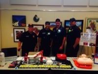 Four uniformed men stand behind a table with medical equipment. A sign says 