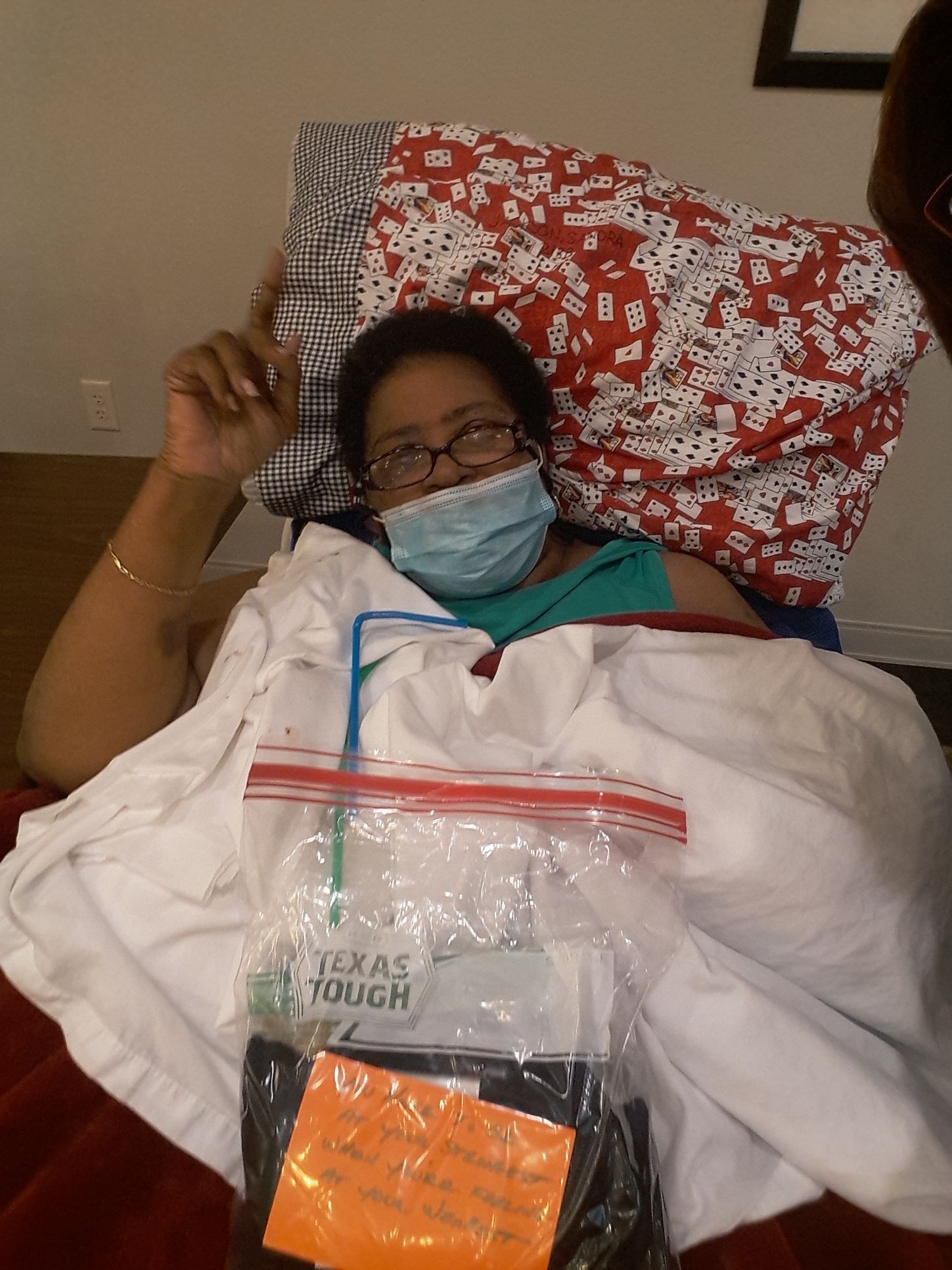 Woman in bed wearing a mask, pointing up. A bag with supplies is visible.