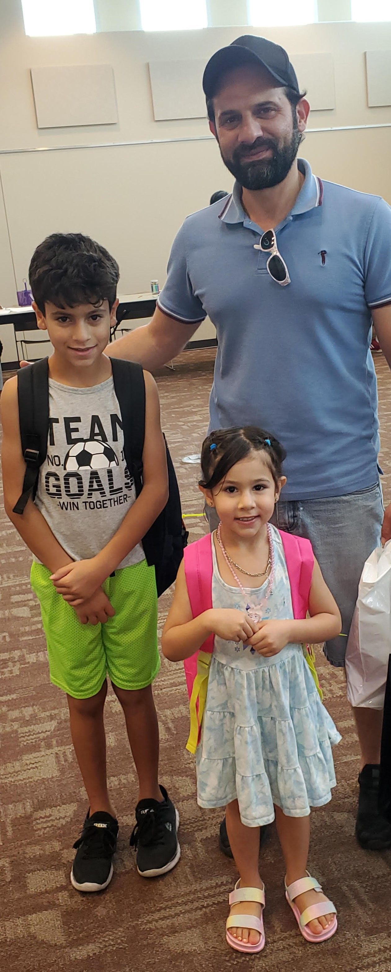 A man and two children pose. The boy wears green shorts and a backpack; the girl, a dress and pink backpack.