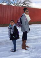Two people standing in snow in front of a red wall. One wears a winter coat, scarf, and boots, the other a winter coat and backpack.