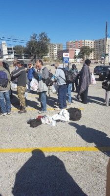 People waiting in a parking lot, some holding bags. A person lies on the ground nearby. Buildings and blue sky in the background.
