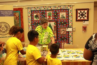 People at a table looking at items. Woman in tie-dye shirt stands behind the table with quilts on display.