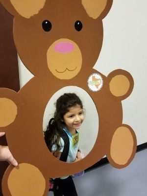 Girl smiling through a cutout in a brown teddy bear; she wears a backpack.