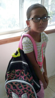 Girl with glasses and a pink backpack stands near a window.