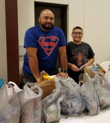 Man in Superman shirt and boy smile, standing behind bags of groceries on a table.