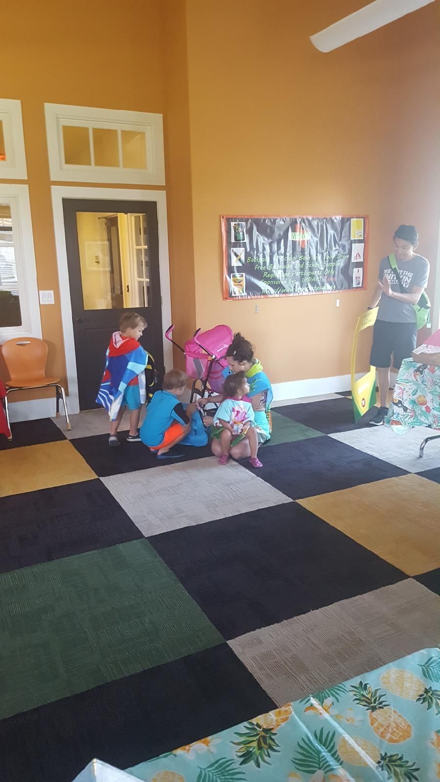 Children playing on a multi-colored rug, near a door and a wall with a poster. A stroller is visible.