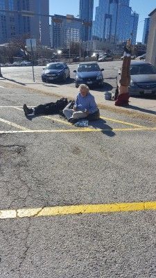Woman sitting in parking lot, reading. Another person lies nearby, upside-down person in background. City skyline.