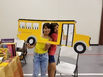 Two girls smiling, posing with a yellow school bus cutout at a book fair.