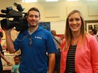 A man holds a video camera on his shoulder next to a woman wearing a pink jacket and smiling.