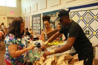 People at a community event, receiving food bags from a volunteer. Quilts hang on the wall.