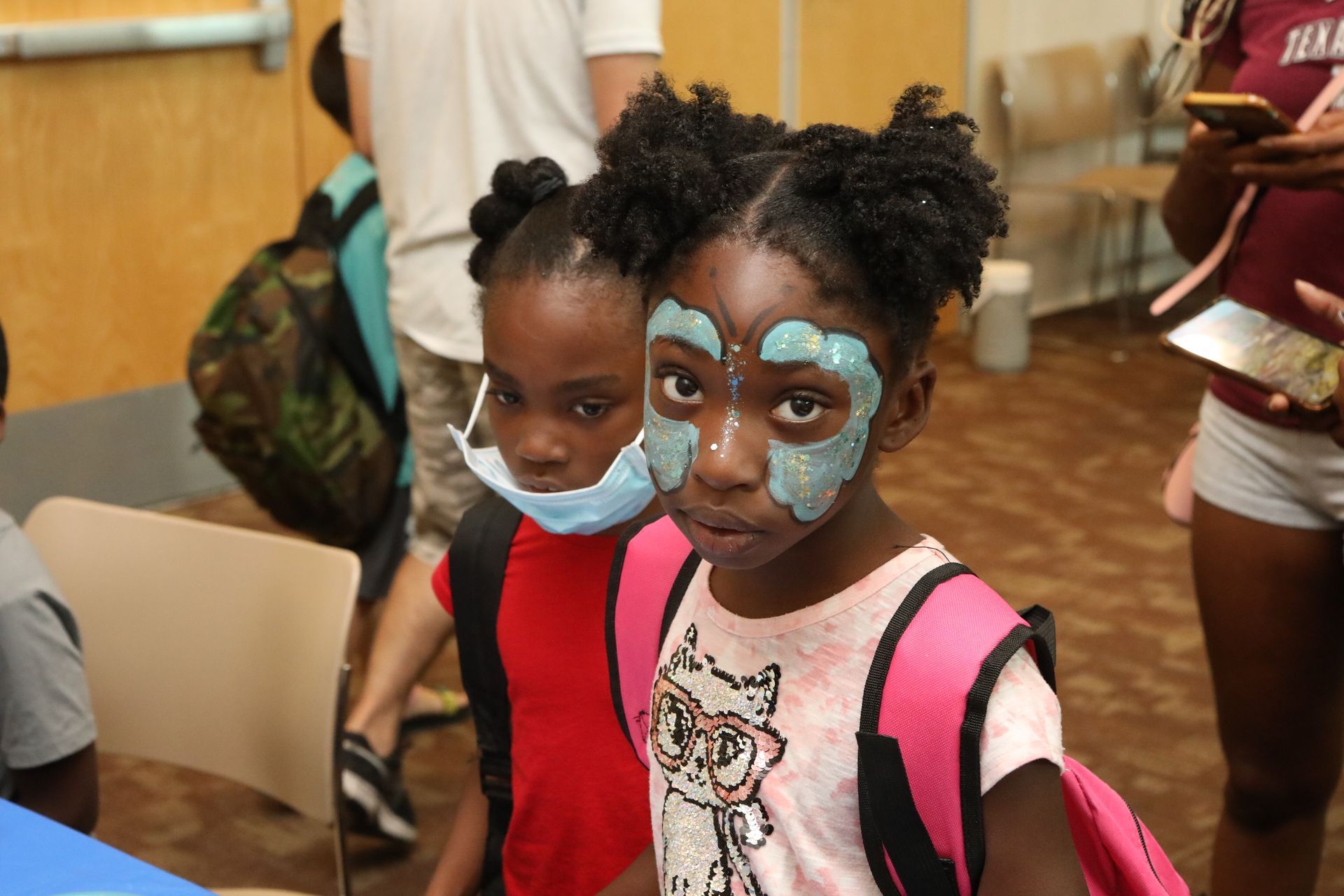 Two young girls with backpacks; one with face paint, the other wearing a mask, inside a room with other people.