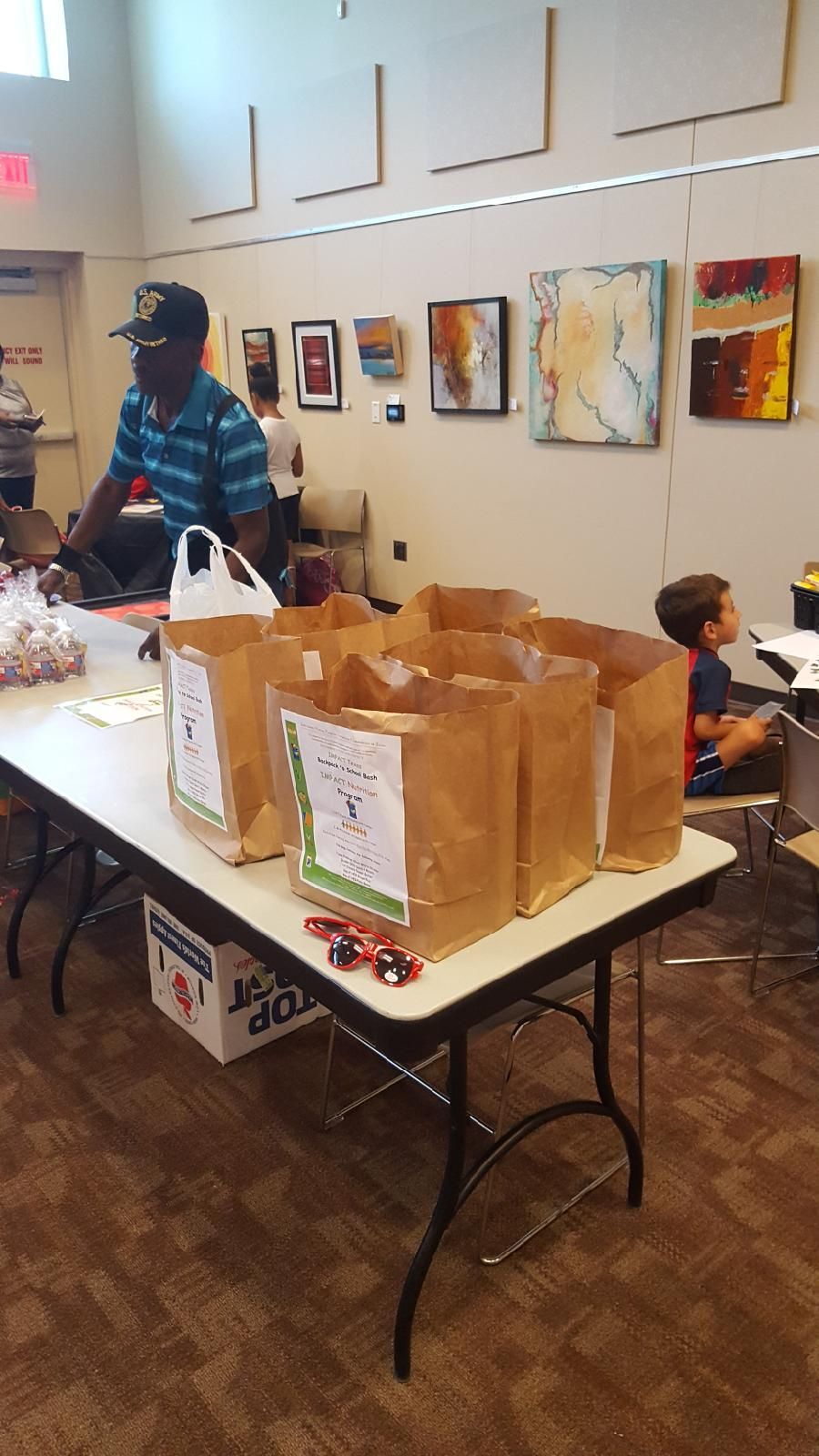 Brown paper bags with labels on a table. A person in a blue shirt and a boy are in the room. Artwork on the wall.