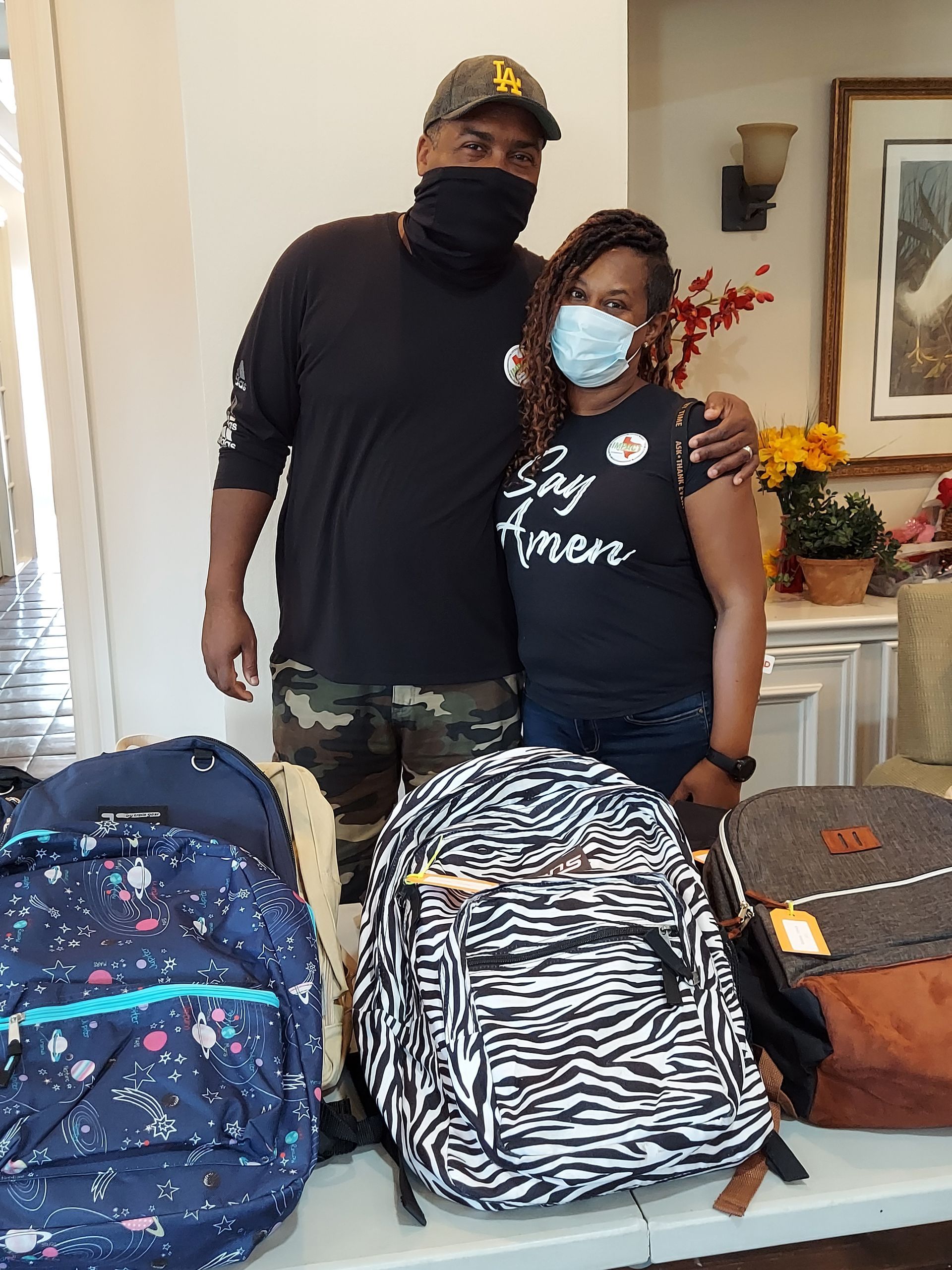 Two people pose with backpacks, possibly for donation. They wear face masks indoors.
