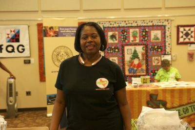 Woman in black shirt stands in front of a table with a decorative quilt backdrop.