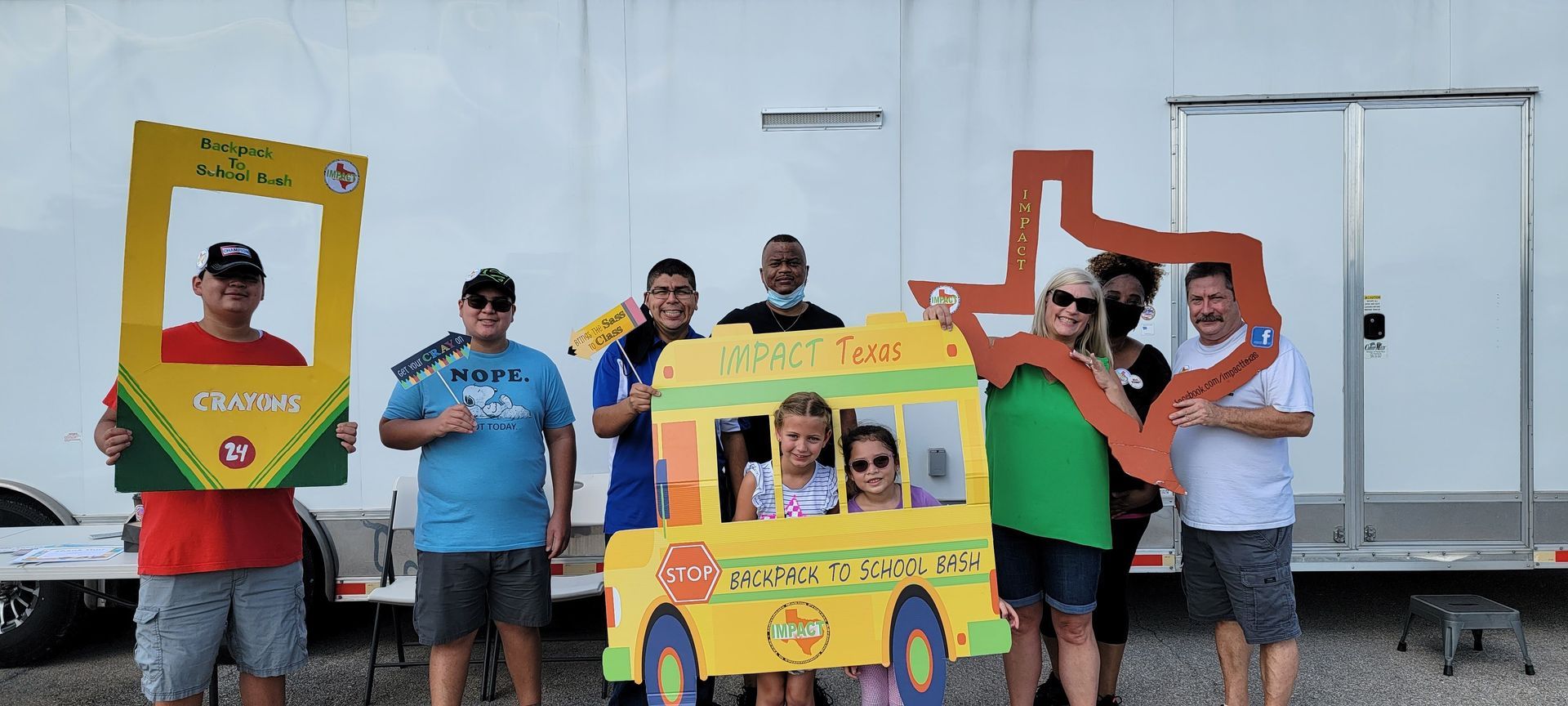 Group of people posing with photo props, including a crayon, school bus, and Texas outline.