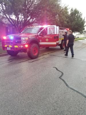 Fire truck with flashing lights parked on a street; firefighters stand nearby, inspecting.