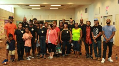Group of people in a brightly lit room. Some wearing aprons, others standing, smiling, or looking at the camera.