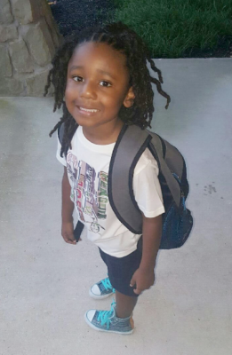 Young child with backpack, smiling, wearing white shirt, shorts, and teal shoes.