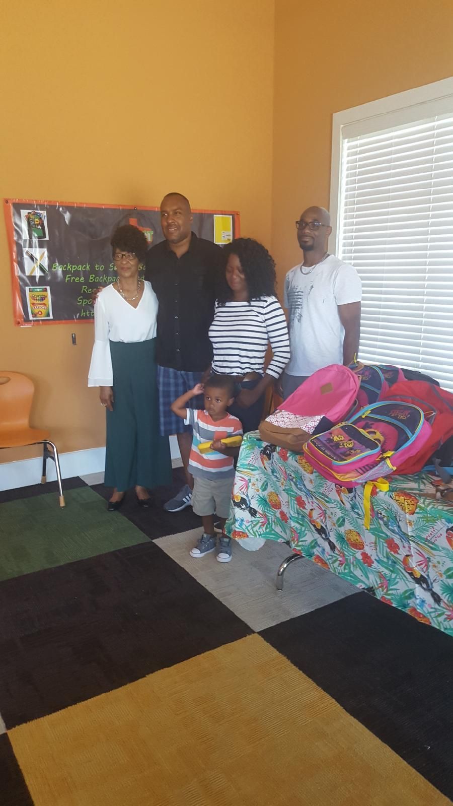 Group of people smiling, posing with backpacks in an orange room. A young child salutes.