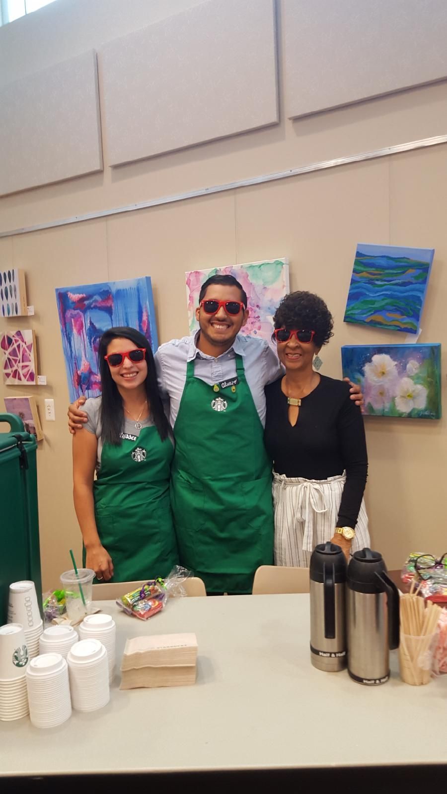 Three people wearing red sunglasses and Starbucks aprons smile behind a counter, with art on the wall.