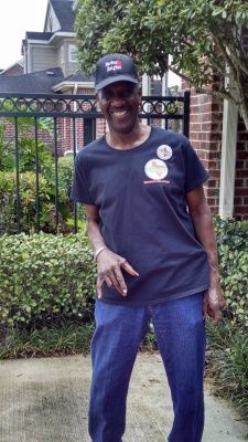 Person in a dark t-shirt and jeans wearing a baseball cap stands outside near a fence and bushes.