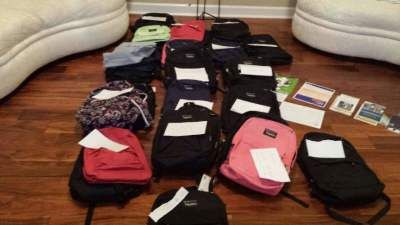 A collection of backpacks, various colors, with white labels, on a wooden floor, near a couch.