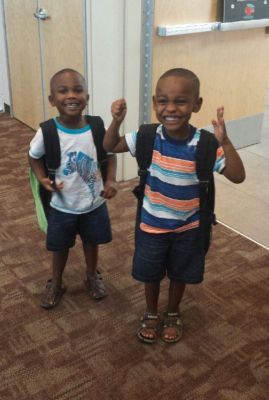 Two young boys with backpacks smile. One raises his arms, in a doorway.