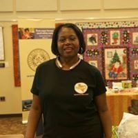 Woman in black shirt smiling, standing indoors, near a banner and a quilt.