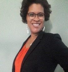 Woman in glasses and blazer smiles, orange top, against a pale green wall.