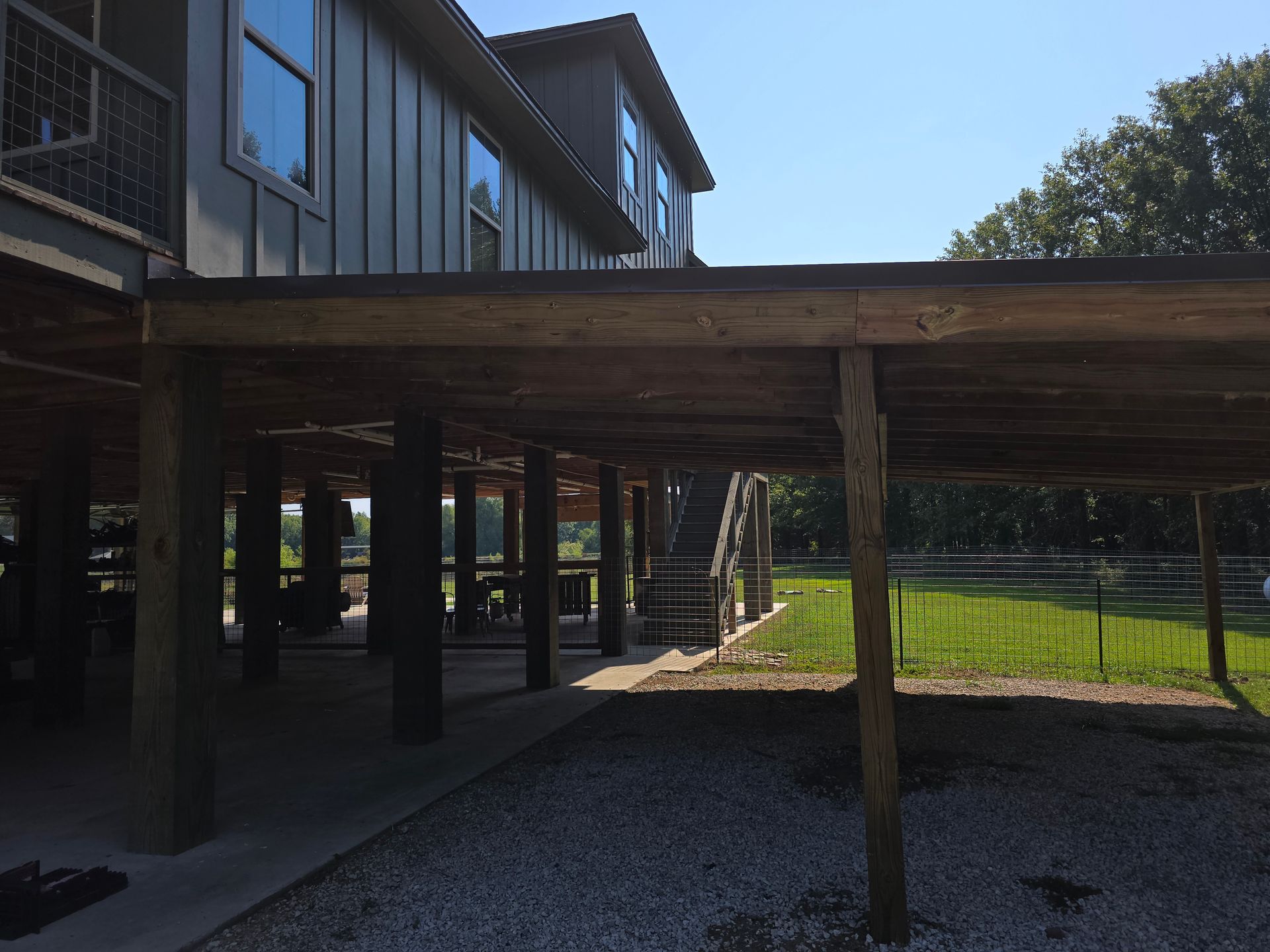 View of a house with a carport underneath. Gray siding, wooden beams, concrete and gravel ground.
