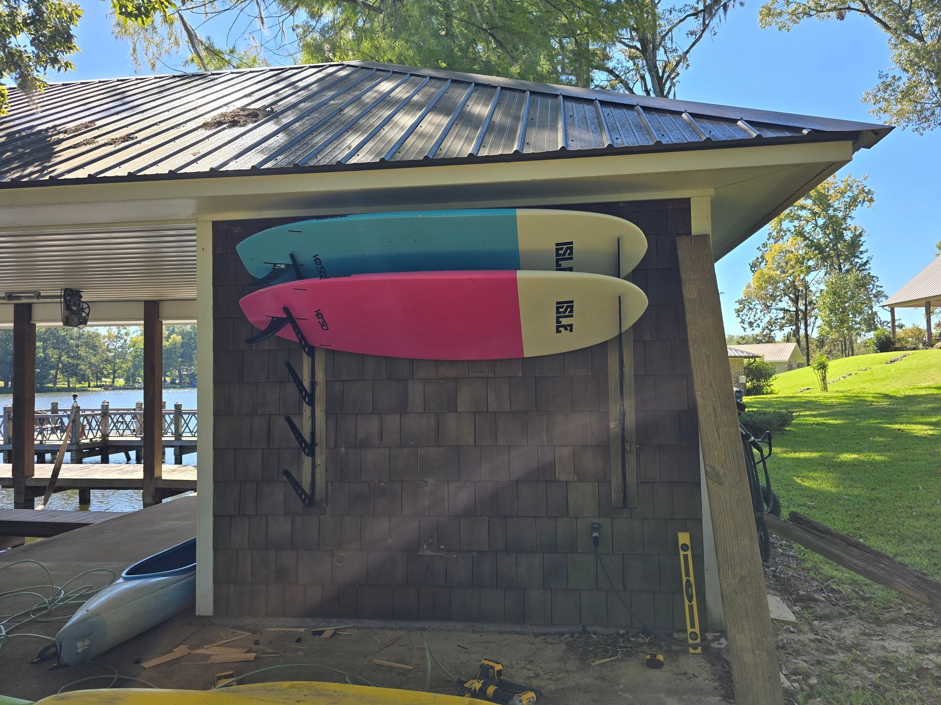 Two paddleboards mounted on a brown shed wall near a lake, under a roof. One is blue/teal, one is pink/red.
