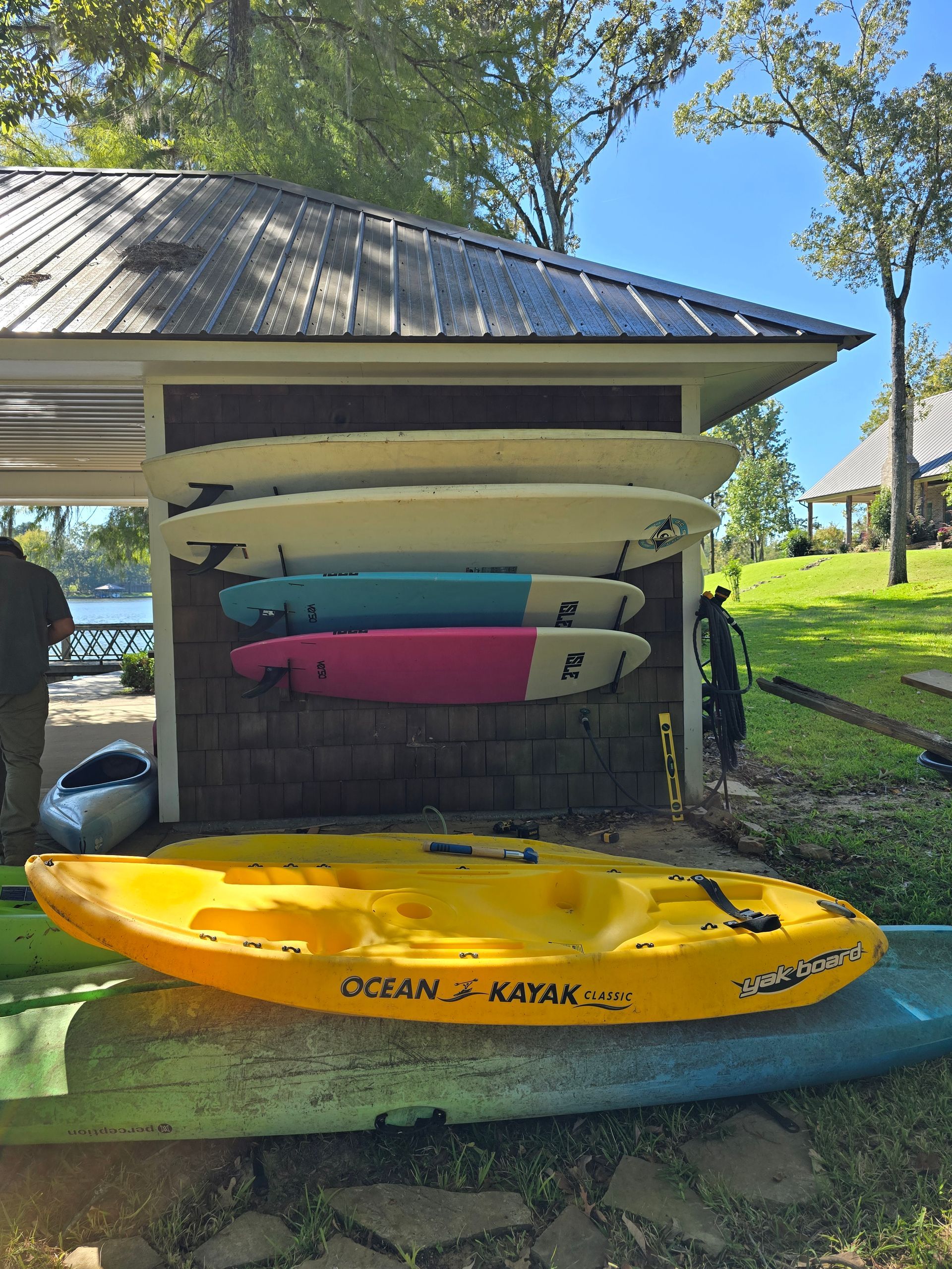 Yellow kayak and three paddleboards on a rack by a building near water.