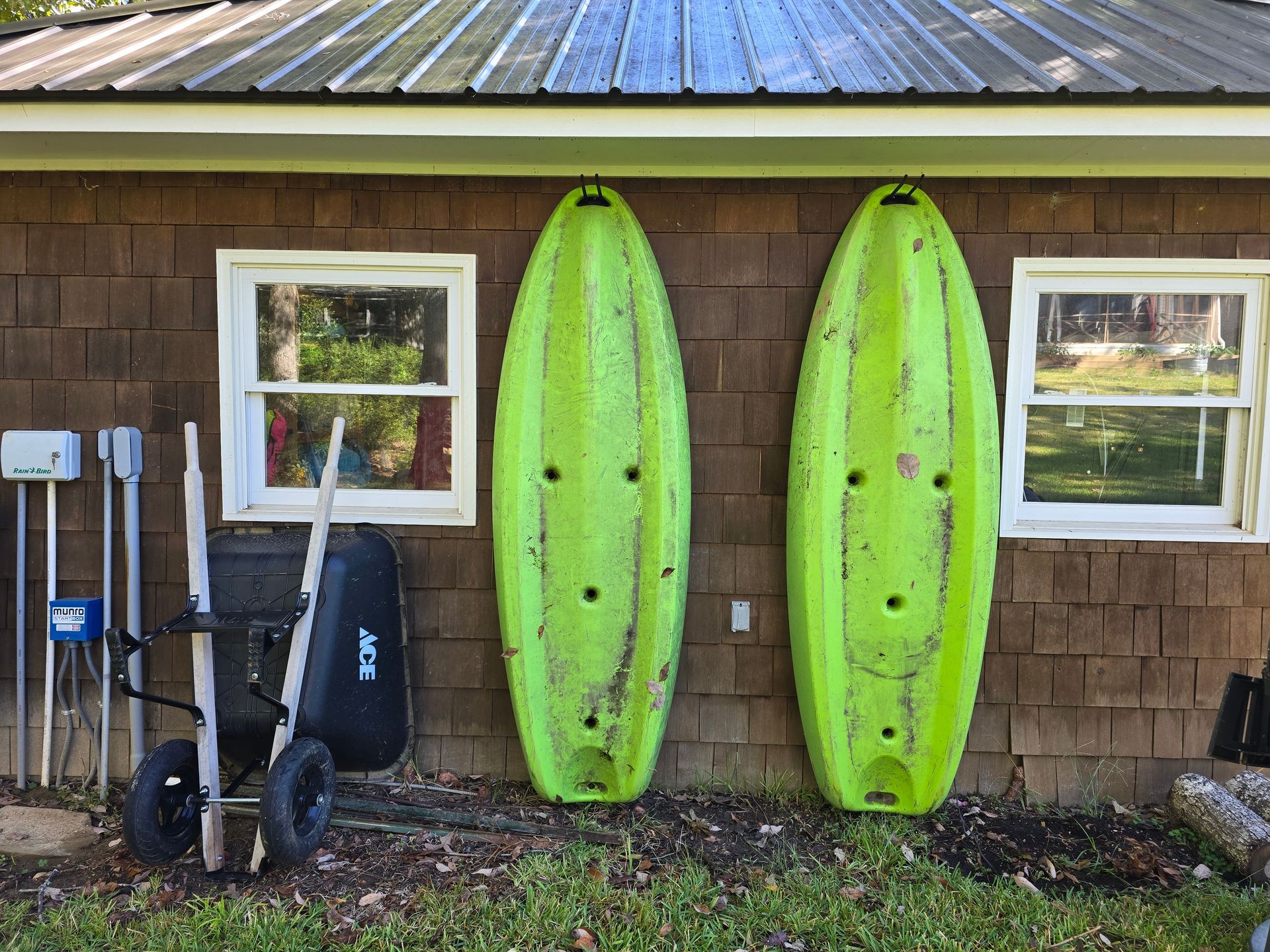 Two green kayaks hanging on a brown shingled wall next to windows; yard tools beside them.
